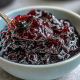 A jar of homemade Black Currant Jelly with a deep purple hue, glistening next to toast points and a silver spreader.