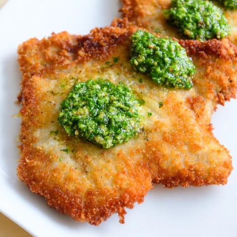 A close-up of a golden, Parmesan-crusted chicken cutlet drizzled with pesto, served alongside lemon wedges for brightness.