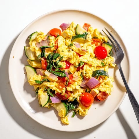 Soft, fluffy scrambled eggs in a pan, dotted with halved cherry tomatoes, fresh spinach, and diced red onions, ready for a quick breakfast.